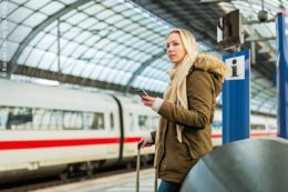 Frau wartet am Bahnhof auf einen Zug und informiert sich über ein Smartphone.